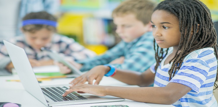 A multi-ethnic group of children are indoors in an elementary school classroom. They are wearing casual clothing. They are working together on a laptop computer together.