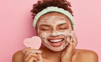 relaxed-joyful-dark-skinned-female-model-washes-face-with-soap-bubbles-enjoys-pampering-session-keeps-eyes-closed-from-pleasure-holds-cosmetic-sponge-cares-about-body-stands-naked-indoor