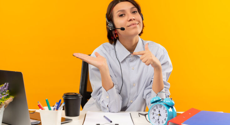 confident-young-call-center-girl-wearing-headset-sitting-desk-showing-empty-hand-pointing-it-isolated-orange